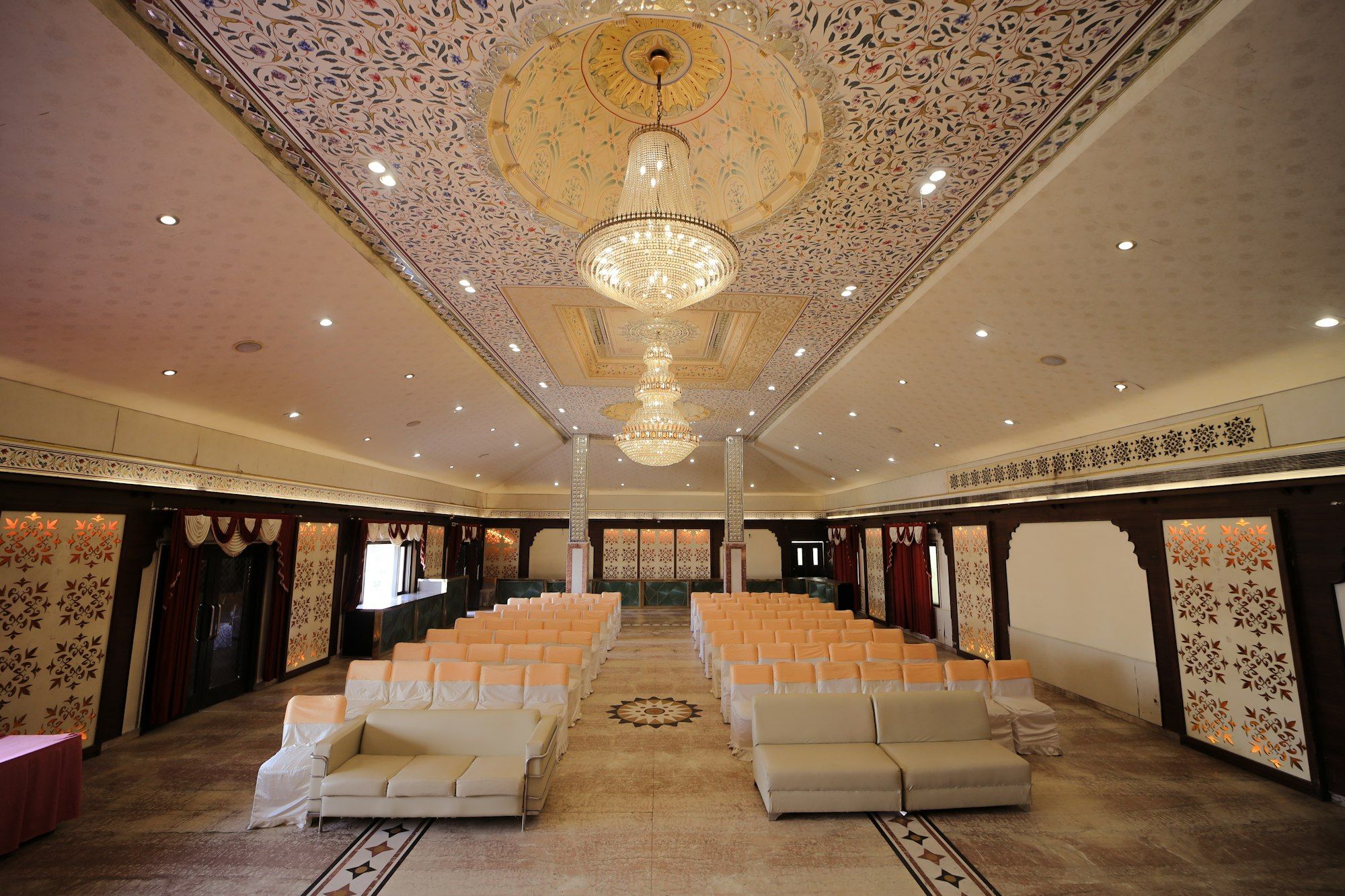 A large banquet hall with a carved white ceiling, two crystal chandeliers and rows of white chairs.