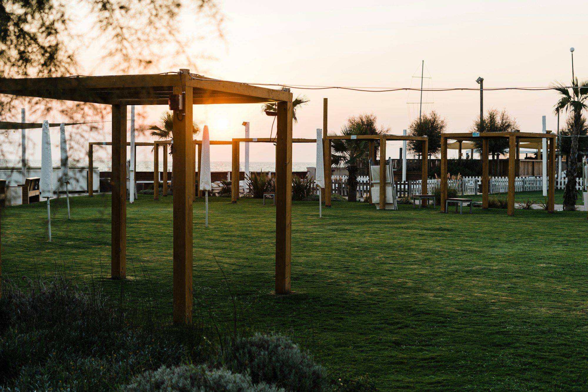 A wide green lawn at golden hour, with timber pergolas and low lights along the edge as the sun sets.