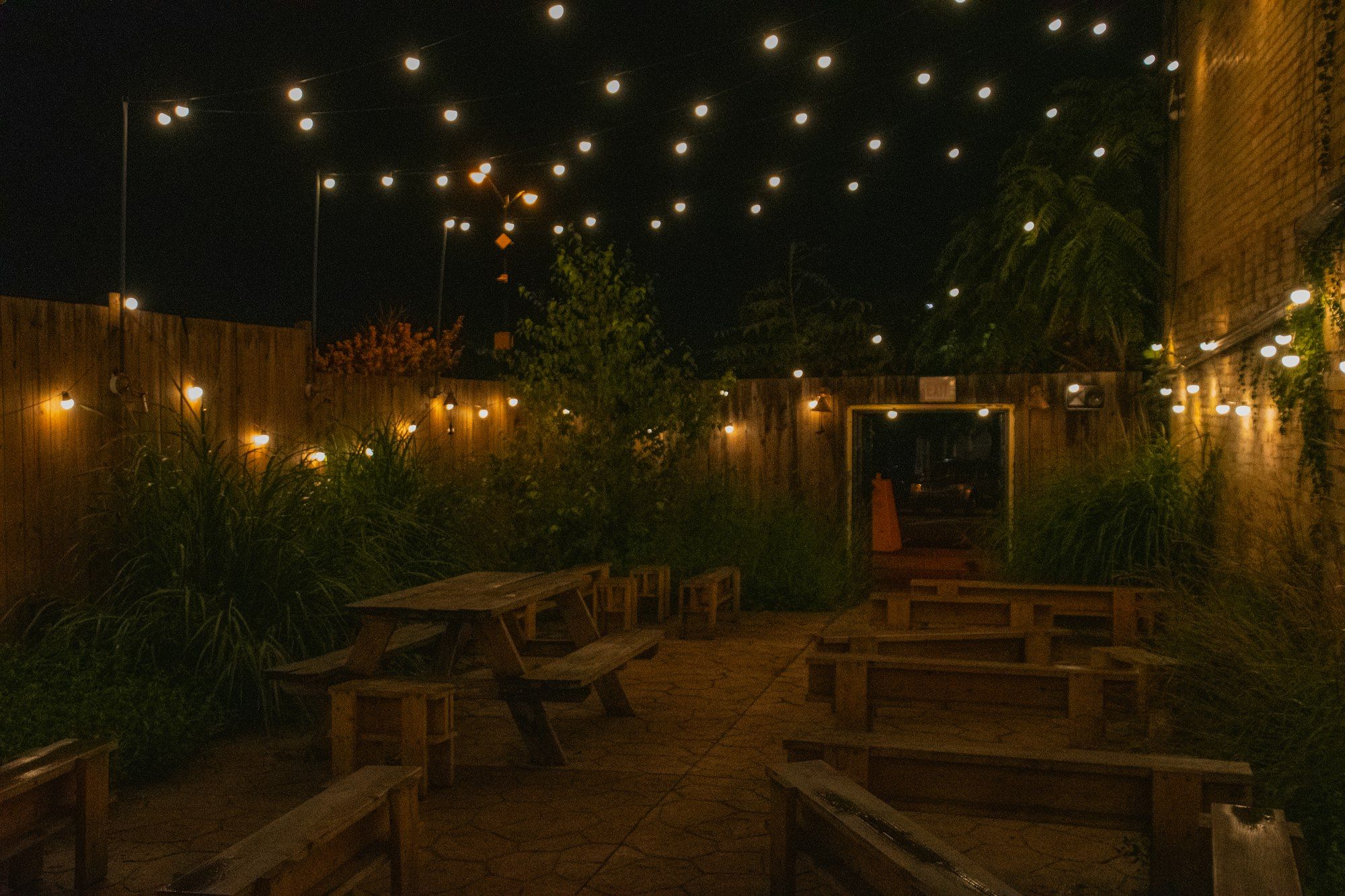 Wooden tables and benches under rows of string lights in an open courtyard at night.